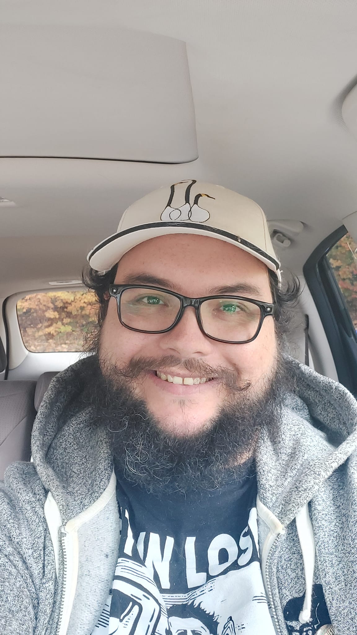 A man with short hair and bushy facial hair sits in a car, smiling